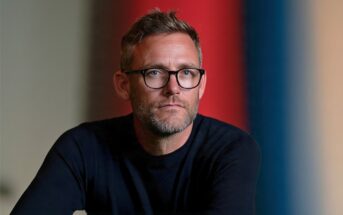 A man with short hair, glasses, and a beard wears a dark sweater and looks at the camera. The background is blurred with vertical red and blue stripes.