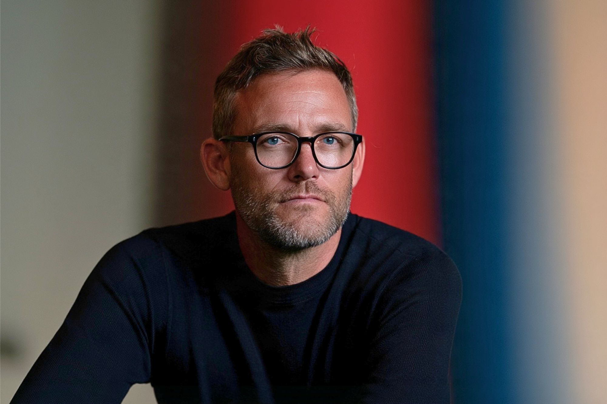 A man with short hair, glasses, and a beard wears a dark sweater and looks at the camera. The background is blurred with vertical red and blue stripes.