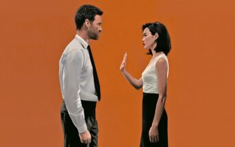 A man in a shirt and tie faces a woman with short dark hair who is holding up her hand to stop him. Both stand against a plain orange background.