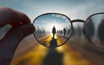 A hand holds eyeglasses with one lens focusing sharply on a person walking down a sunlit street, while the surrounding scene remains blurred and out of focus.