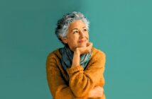 An older woman with short gray hair, wearing a mustard yellow sweater and a blue scarf, smiles thoughtfully while resting her chin on her hand against a teal background.