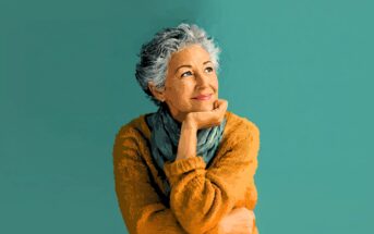 An older woman with short gray hair, wearing a mustard yellow sweater and a blue scarf, smiles thoughtfully while resting her chin on her hand against a teal background.