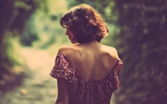 A woman with short brown hair in a floral off-shoulder dress stands on a sunlit path, facing away and looking over her shoulder. The background is blurred with greenery, creating a soft, dreamy atmosphere.