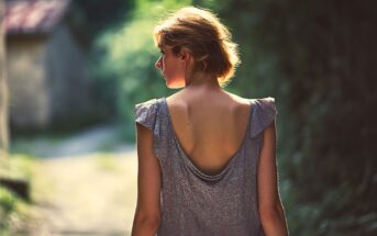 A woman with short hair wearing a backless dress walks outdoors on a sunlit path, surrounded by greenery, with her back turned to the camera.