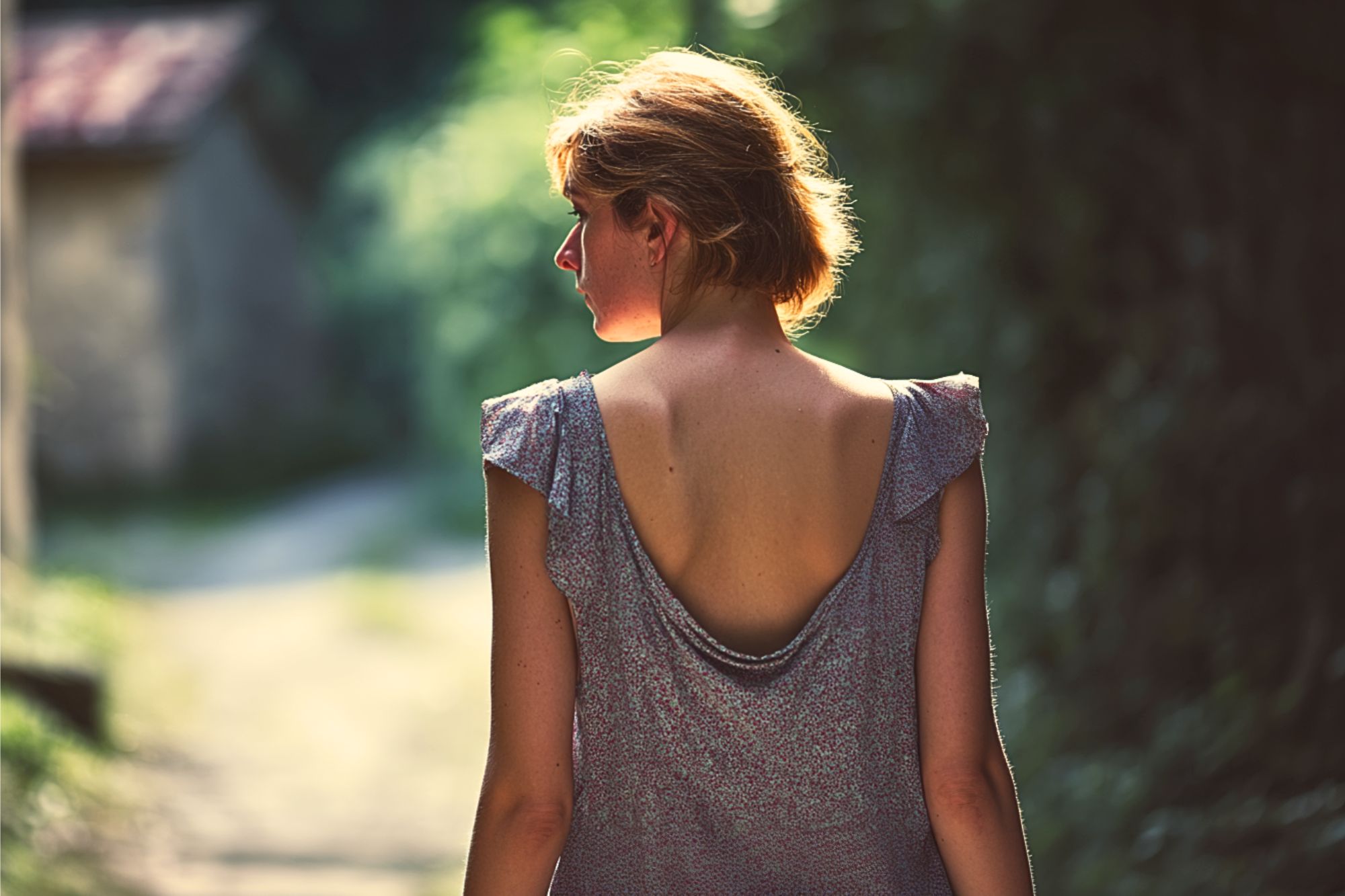A woman with short hair wearing a backless dress walks outdoors on a sunlit path, surrounded by greenery, with her back turned to the camera.
