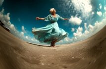 An older woman in a flowing blue dress joyfully dances barefoot on a sandy beach under a dramatic, cloudy sky, captured with a fisheye lens that curves the horizon.