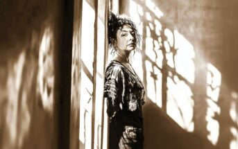 A person with curly hair leans against a wall in a narrow hallway, illuminated by dramatic sunlight and shadows streaming through a patterned window. The scene is in sepia tones, giving it a vintage feel.