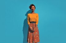 A woman stands against a bright blue background, eyes closed and smiling gently. She wears a yellow top, an orange patterned skirt, hoop earrings, and a necklace, with her hands loosely clasped in front of her.