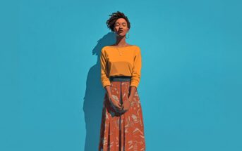 A woman stands against a bright blue background, eyes closed and smiling gently. She wears a yellow top, an orange patterned skirt, hoop earrings, and a necklace, with her hands loosely clasped in front of her.