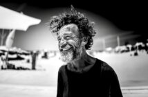 A man with curly hair and a beard smiles broadly while standing outdoors on a sunny beach. He wears a dark shirt and appears joyful. The background is blurred with beach umbrellas and people. The photo is in black and white.