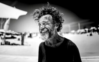 A man with curly hair and a beard smiles broadly while standing outdoors on a sunny beach. He wears a dark shirt and appears joyful. The background is blurred with beach umbrellas and people. The photo is in black and white.