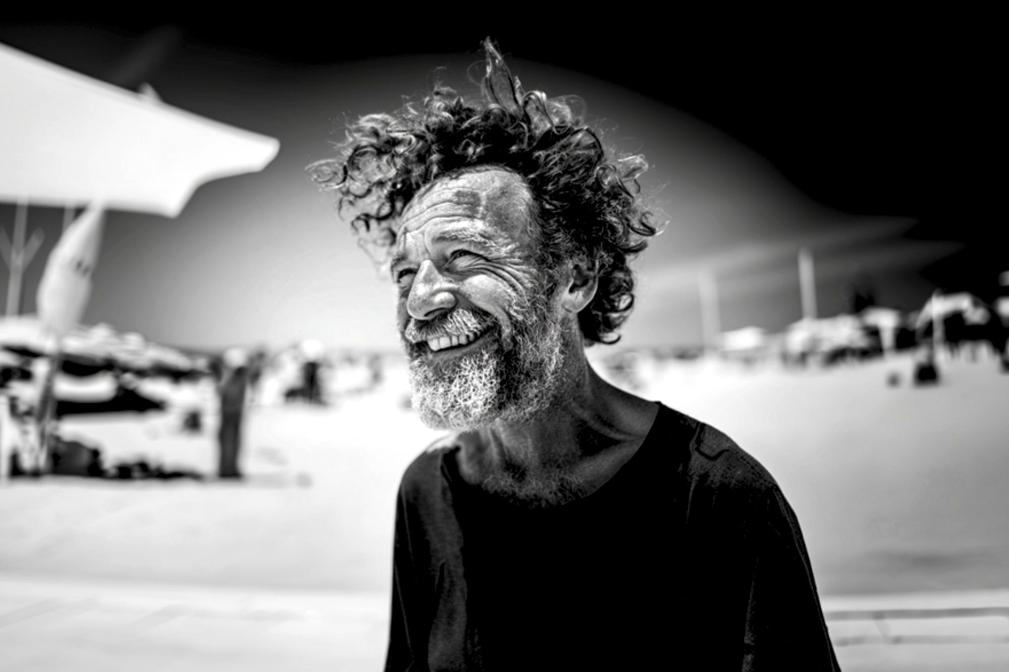 A man with curly hair and a beard smiles broadly while standing outdoors on a sunny beach. He wears a dark shirt and appears joyful. The background is blurred with beach umbrellas and people. The photo is in black and white.