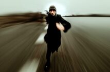 A person wearing dark clothing and a beanie is running toward the camera on a blurred road, creating a dynamic sense of speed and motion. The background appears empty and overcast.