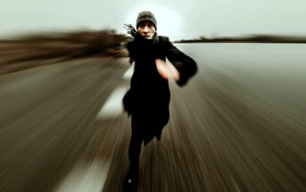 A person wearing dark clothing and a beanie is running toward the camera on a blurred road, creating a dynamic sense of speed and motion. The background appears empty and overcast.