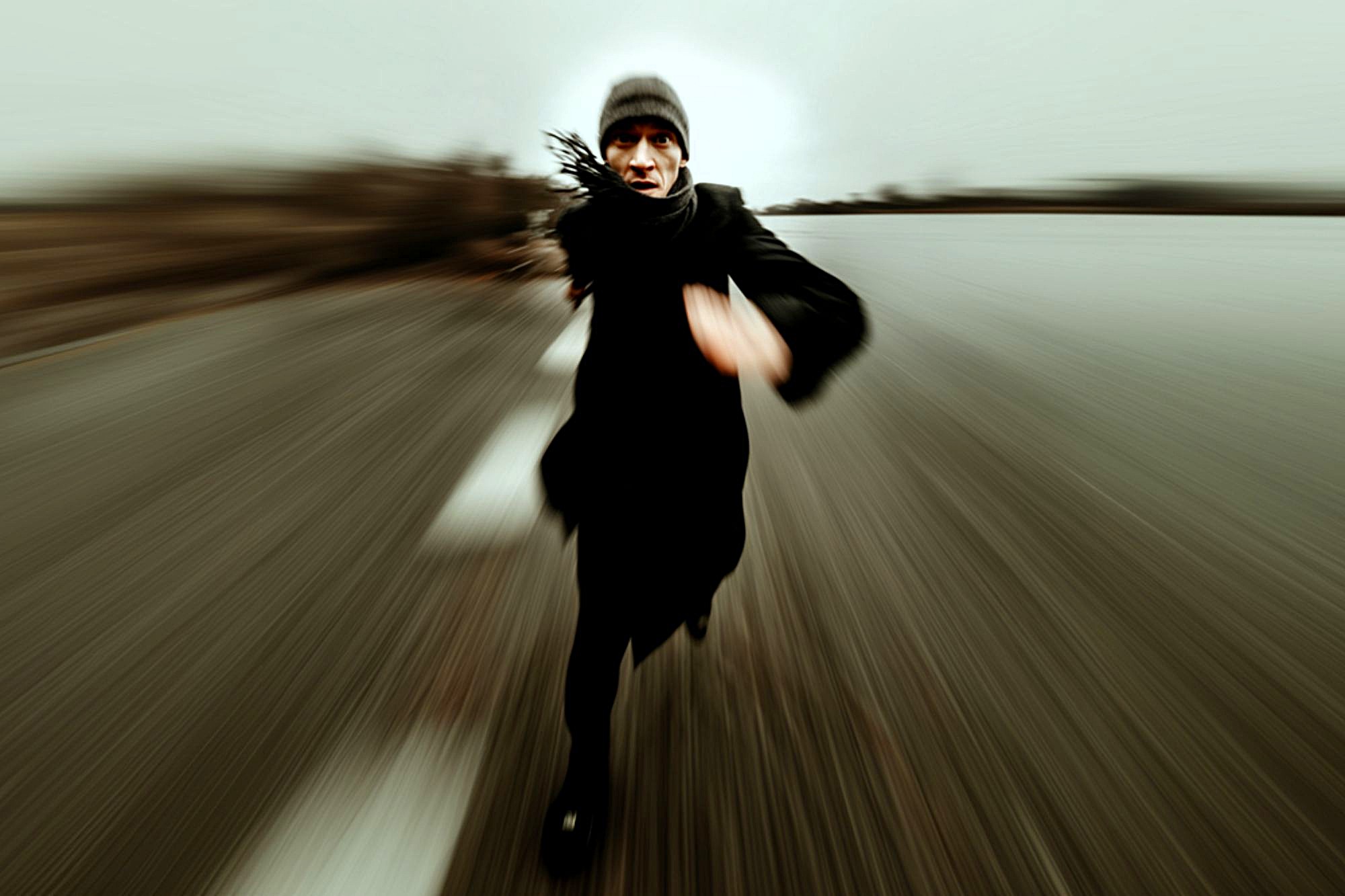 A person wearing dark clothing and a beanie is running toward the camera on a blurred road, creating a dynamic sense of speed and motion. The background appears empty and overcast.