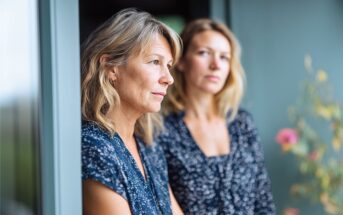 Two women with similar features, both wearing blue patterned shirts, stand side by side near a window. One looks thoughtfully into the distance while the other gazes slightly past the camera.