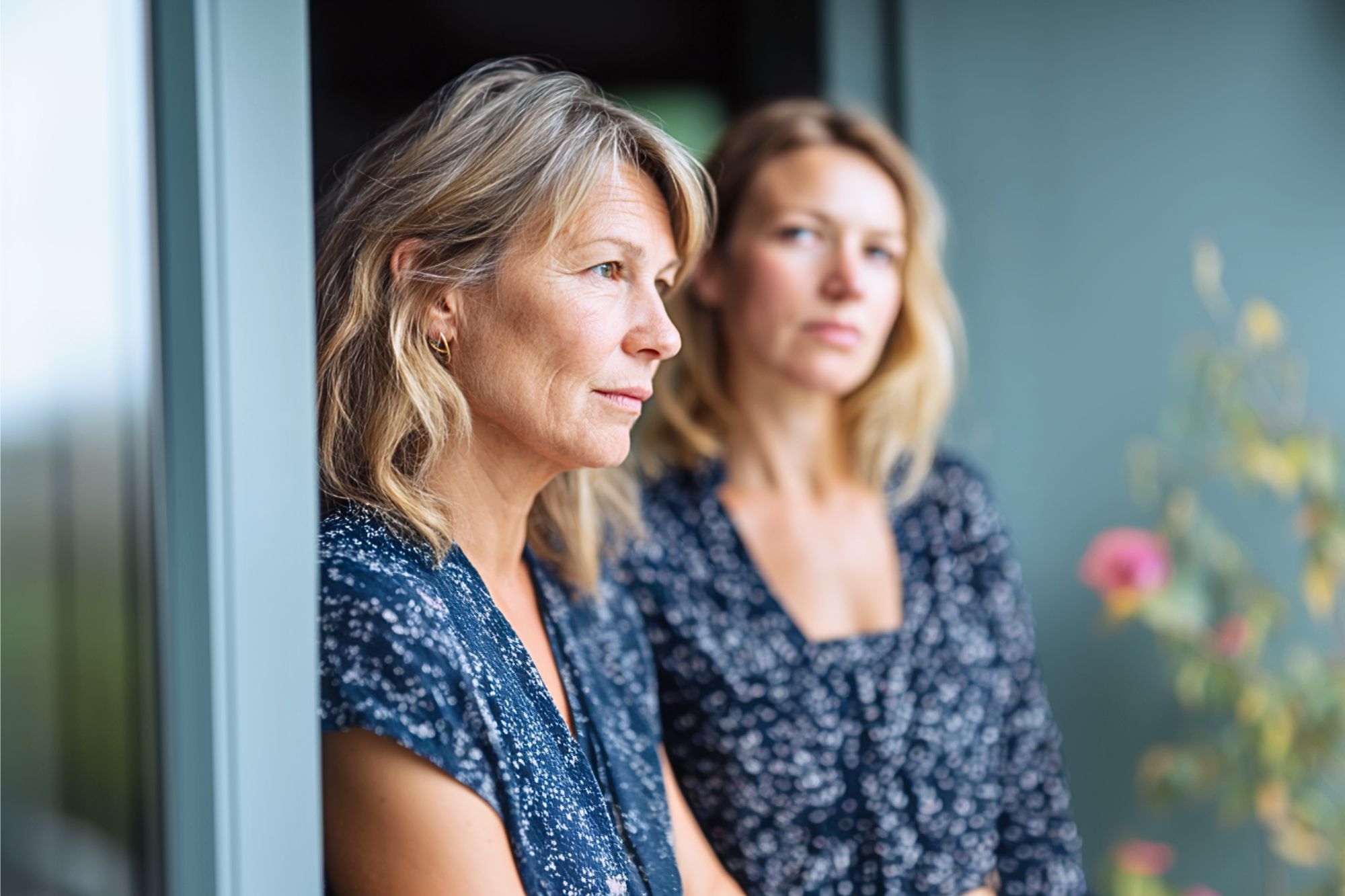 Two women with similar features, both wearing blue patterned shirts, stand side by side near a window. One looks thoughtfully into the distance while the other gazes slightly past the camera.