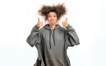 A woman with messy hair wearing an oversized hoodie gives two thumbs up while making a sad, pouty face against a white background.