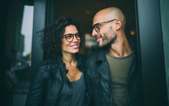 A smiling woman and man, both wearing glasses and black jackets, stand closely together in a doorway, looking at each other warmly. The background is blurred, drawing focus to their faces and expressions.