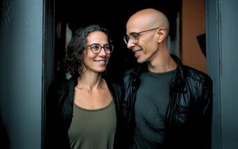 A smiling woman and man stand close together in a doorway, both wearing glasses. The woman has curly hair and wears a green tank top; the man is bald, wears a black jacket, and looks at her warmly.