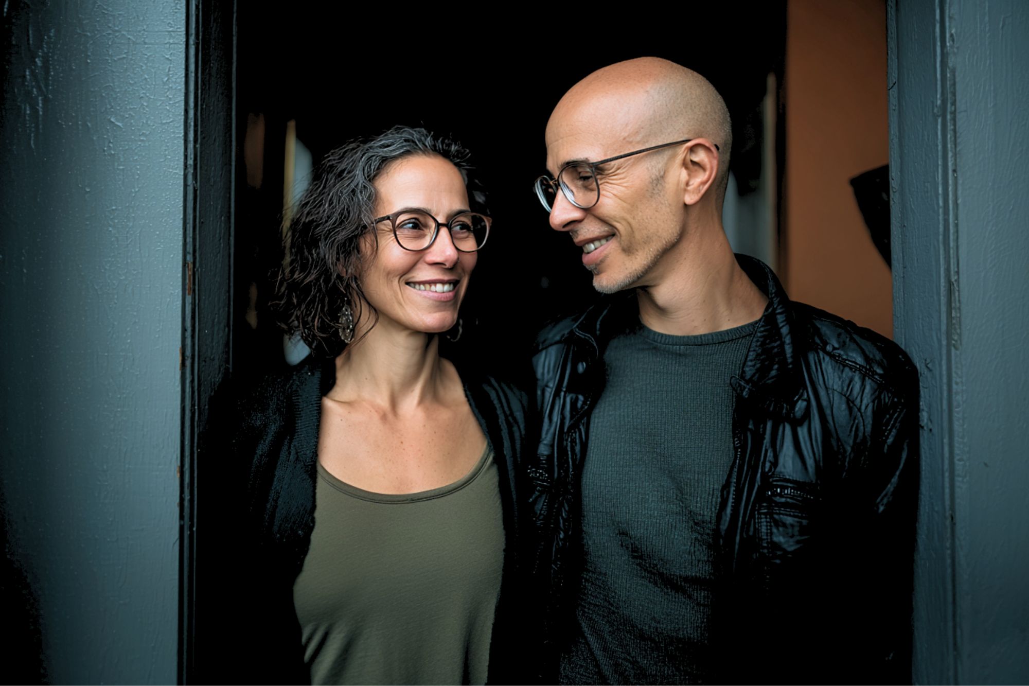 A smiling woman and man stand close together in a doorway, both wearing glasses. The woman has curly hair and wears a green tank top; the man is bald, wears a black jacket, and looks at her warmly.