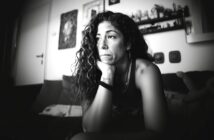 A woman with long curly hair sits on a couch, resting her chin on her hand and gazing thoughtfully into the distance in a softly lit, monochrome room with shelves and framed art on the wall behind her.