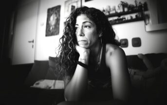 A woman with long curly hair sits on a couch, resting her chin on her hand and gazing thoughtfully into the distance in a softly lit, monochrome room with shelves and framed art on the wall behind her.