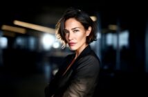 A woman with short brown hair and a confident expression stands with arms crossed, wearing a dark blazer in a dimly lit, modern office with sunlight shining behind her.