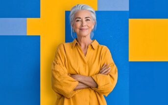 A smiling older woman with gray hair and a yellow shirt stands with arms crossed in front of a bold geometric background of yellow and blue rectangles.
