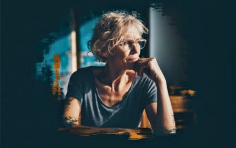 An older woman with curly gray hair and glasses looks thoughtfully out of a window, resting her chin on her hand. She wears a gray shirt, and the image has a dark, artistic brushed border effect.
