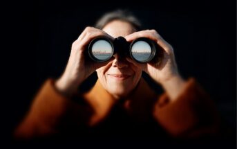 An older woman in a brown coat smiles while looking through binoculars. The reflection in the binocular lenses shows a city skyline across a body of water. The background is dark, highlighting her face.