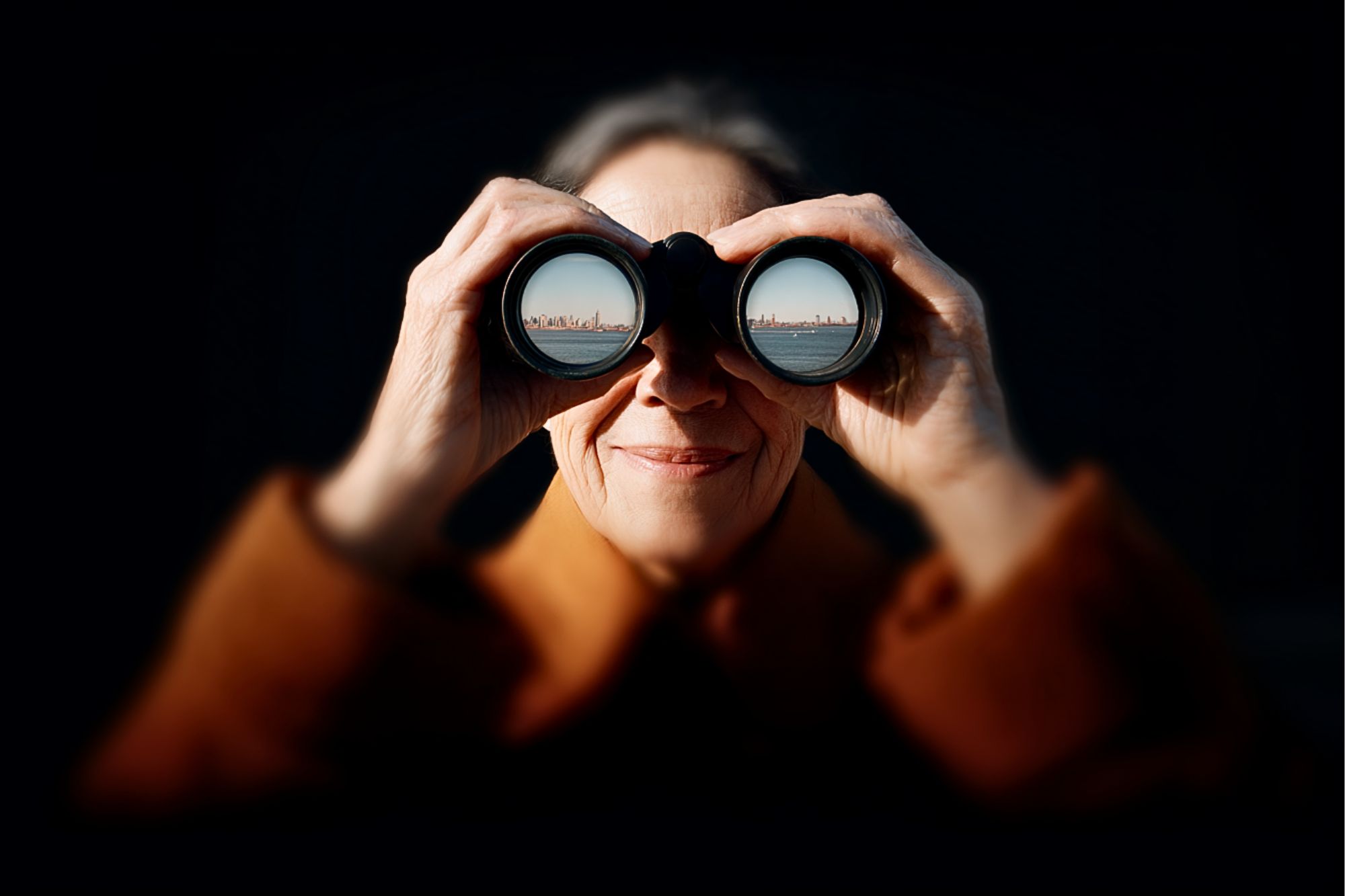 An older woman in a brown coat smiles while looking through binoculars. The reflection in the binocular lenses shows a city skyline across a body of water. The background is dark, highlighting her face.