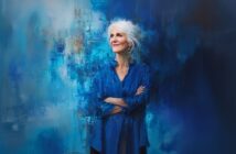 An older woman with white hair and a blue blouse stands with arms crossed, smiling slightly, against an abstract, painterly blue background.