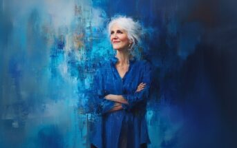 An older woman with white hair and a blue blouse stands with arms crossed, smiling slightly, against an abstract, painterly blue background.