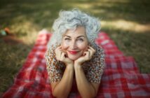 An older woman with curly gray hair smiles while lying on a red checkered picnic blanket outdoors, resting her chin on her hands and looking up at the camera. Grass and sunlight are visible in the background.