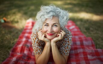 An older woman with curly gray hair smiles while lying on a red checkered picnic blanket outdoors, resting her chin on her hands and looking up at the camera. Grass and sunlight are visible in the background.