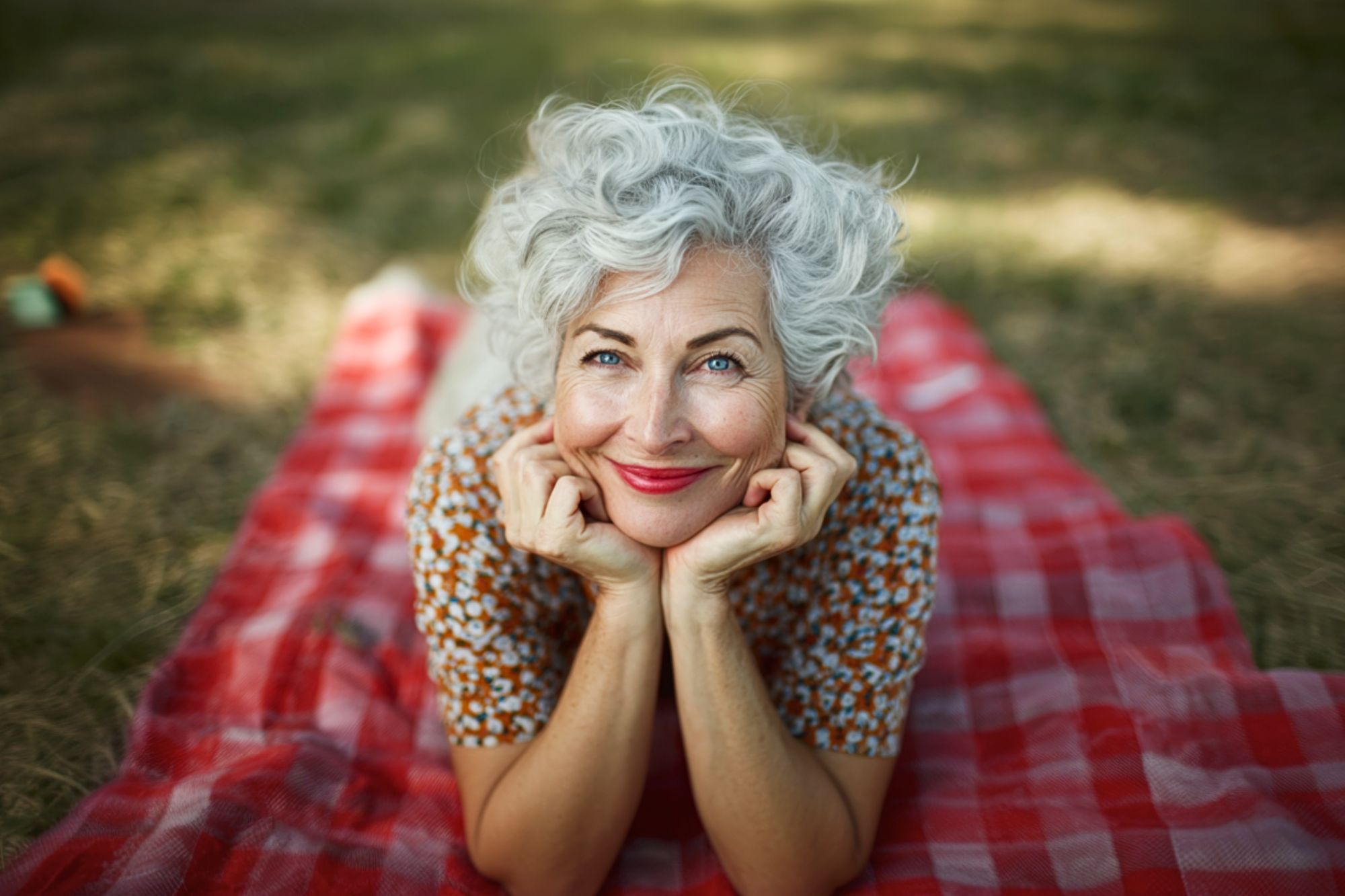 An older woman with curly gray hair smiles while lying on a red checkered picnic blanket outdoors, resting her chin on her hands and looking up at the camera. Grass and sunlight are visible in the background.