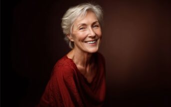 An older woman with short gray hair smiles warmly while wearing a textured, deep red top. The background is a plain dark brown, creating a soft and elegant portrait.