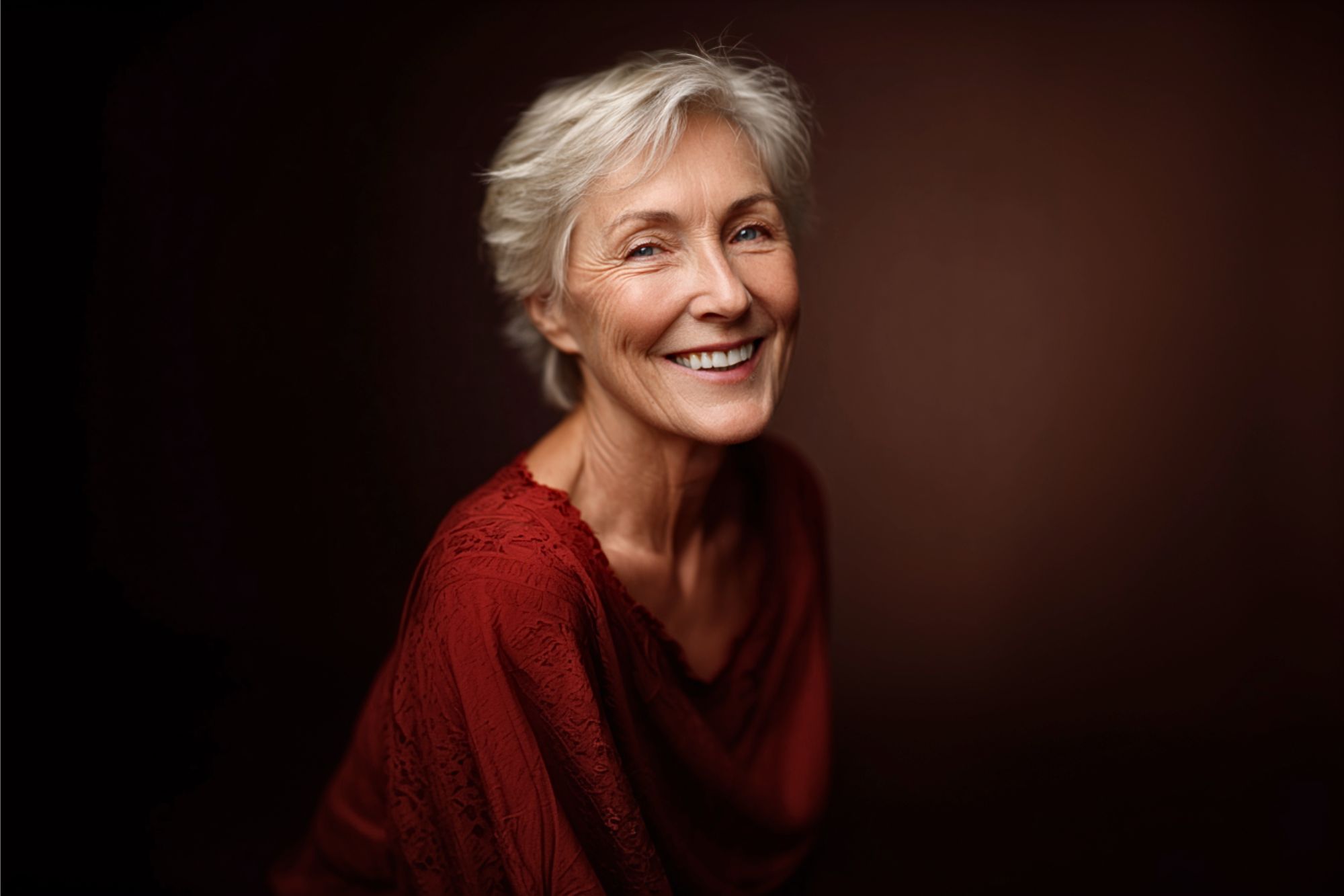 An older woman with short gray hair smiles warmly while wearing a textured, deep red top. The background is a plain dark brown, creating a soft and elegant portrait.
