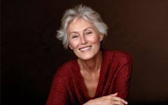 Smiling older woman with short gray hair wearing a textured red top, sitting with her arms crossed against a plain dark background.
