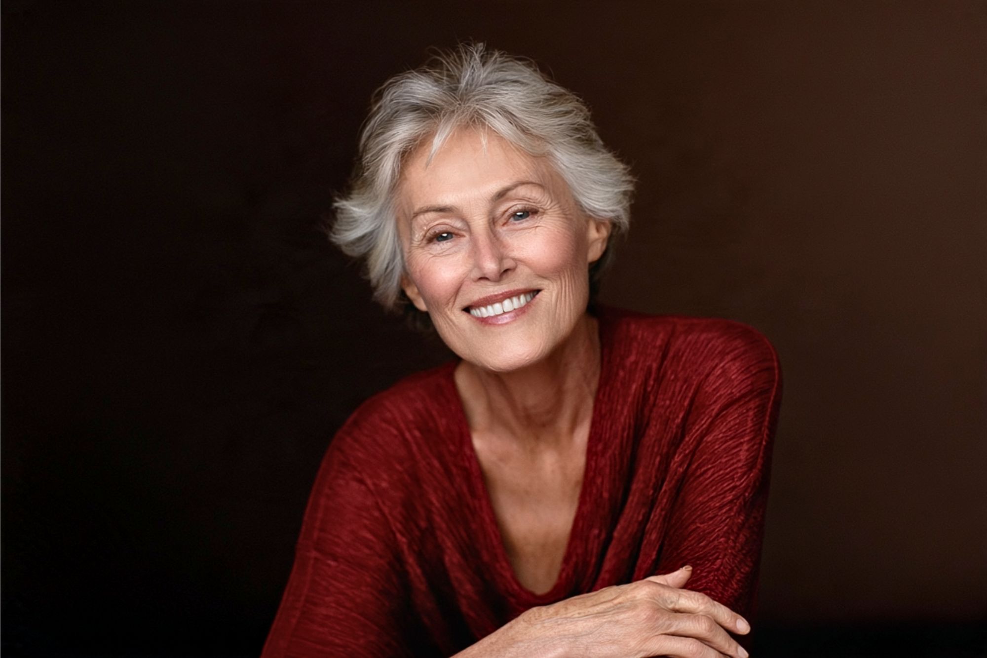 Smiling older woman with short gray hair wearing a textured red top, sitting with her arms crossed against a plain dark background.