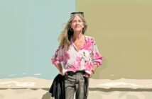 A woman with long blonde hair stands smiling in front of a split pastel background, wearing a pink floral blouse, gray pants, and sunglasses on her head. Her hands are in her pockets.