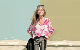 A woman with long blonde hair stands smiling in front of a split pastel background, wearing a pink floral blouse, gray pants, and sunglasses on her head. Her hands are in her pockets.