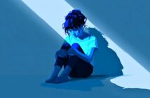 A woman with dark hair sits barefoot on the floor in a mostly dark room, illuminated by a beam of light from above. She looks down, hugging her knees, creating a contemplative and somber mood.