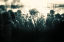 A woman stands alone in a crowd, looking down with a somber expression. The image is in black and white with a soft, blurry focus, creating a moody and introspective atmosphere.