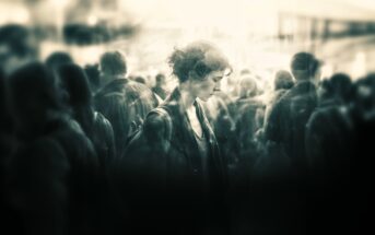 A woman stands alone in a crowd, looking down with a somber expression. The image is in black and white with a soft, blurry focus, creating a moody and introspective atmosphere.