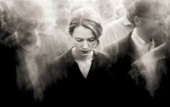 A woman stands with her head bowed amid a blurred, crowded group of people, creating a sense of isolation and introspection despite the busy surroundings. The image is in black and white.