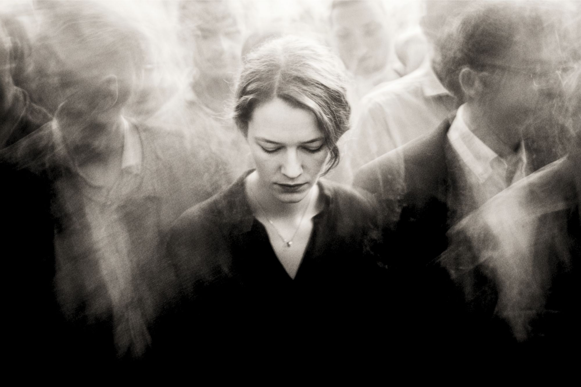 A woman stands with her head bowed amid a blurred, crowded group of people, creating a sense of isolation and introspection despite the busy surroundings. The image is in black and white.