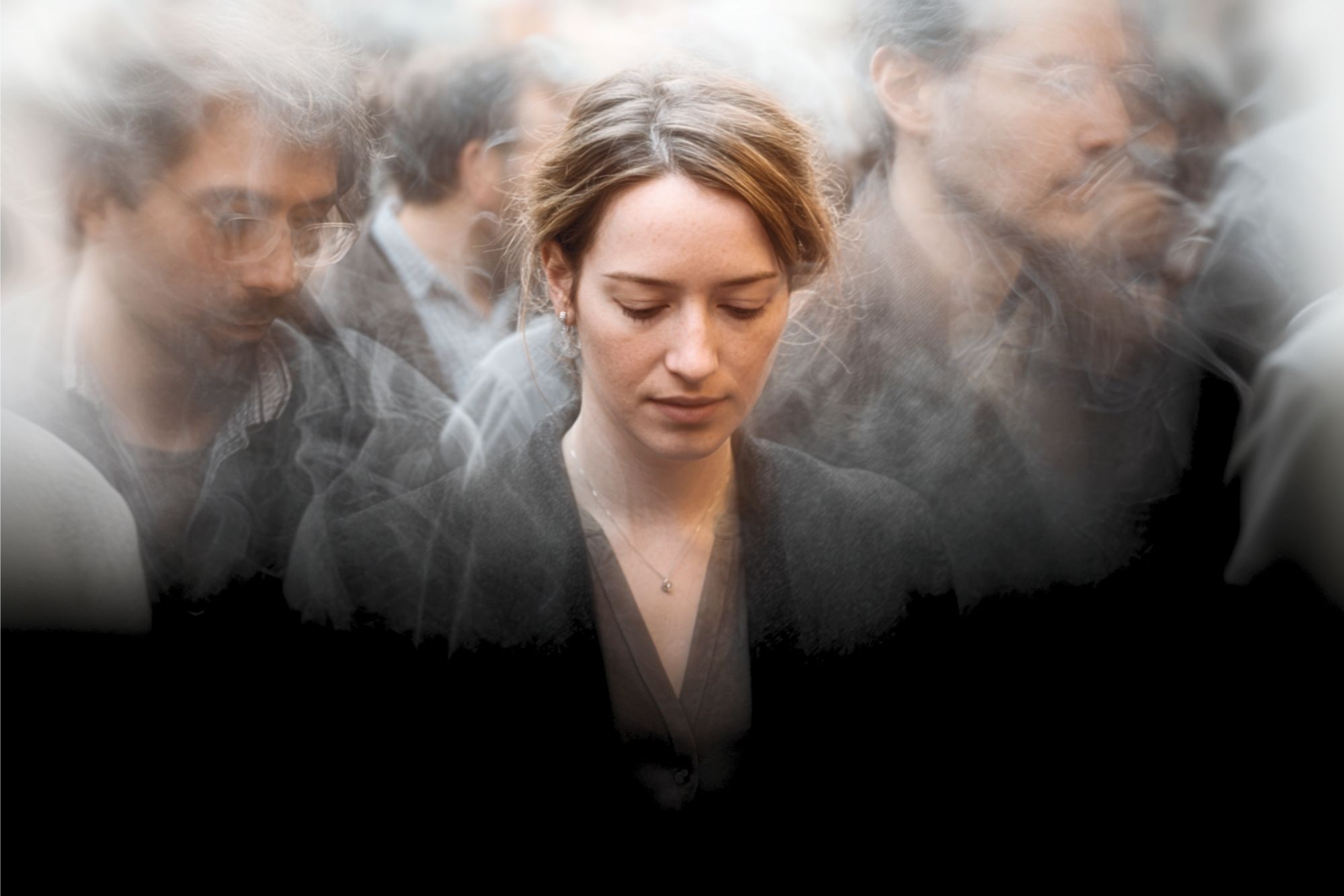 A woman stands with her head bowed, eyes closed, surrounded by blurred, indistinct figures in a crowd, creating an effect of isolation and introspection amidst others.