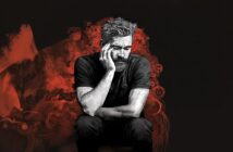 A man with a beard sits with his head resting on his hand, appearing deep in thought or sadness. The background features abstract red and black artistic elements surrounding him.